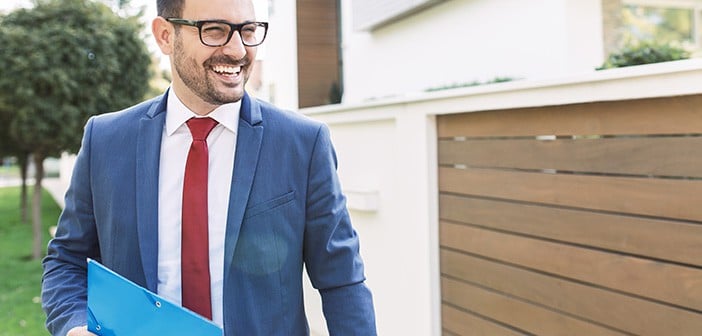 smiling brown-haired man with glasses and shadow of a beard evaluates exterior of a residential property while wearing suit and carrying a folder