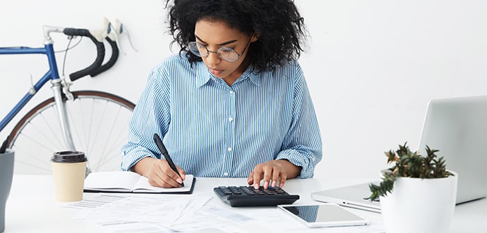 businesswoman doing paperwork at office desk, working through finances, using calculator and making notes in her notebook with pen