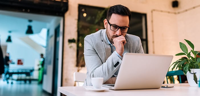 Pensive young entrepreneur looking at laptop screen and drinking coffee at table in cafe