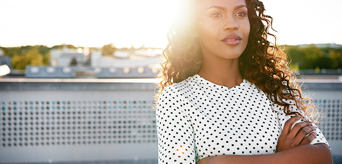 Confident young woman standing on an urban rooftop daydreaming with her arms folded backlit by the bright flare of the rising sun