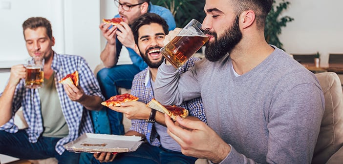 Group of four male friends drinking beer and eating pizza at home.