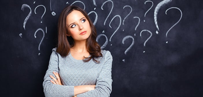 young dark-haired woman in gray sweater standing in front of blackboard with questions drawn on it in white chalk