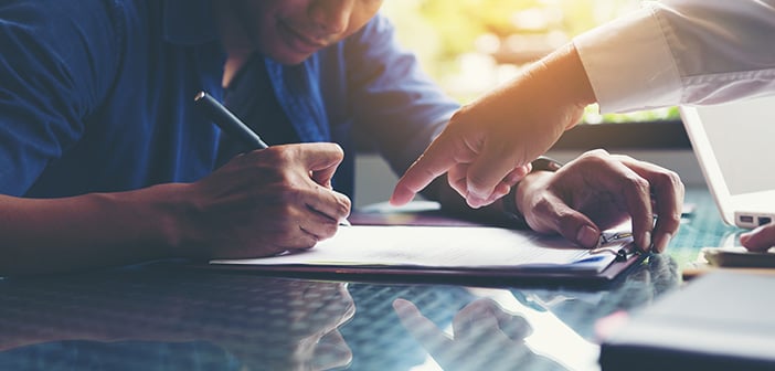 Person sitting at a desk signing paperwork with guidance from another person who is pointing at a line item