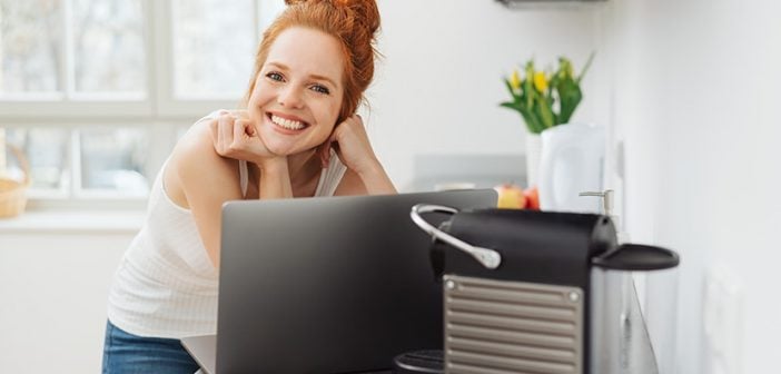 Young woman relaxing in a kitchen with a laptop computer balanced on the counter looking over the top to smile happily at camera