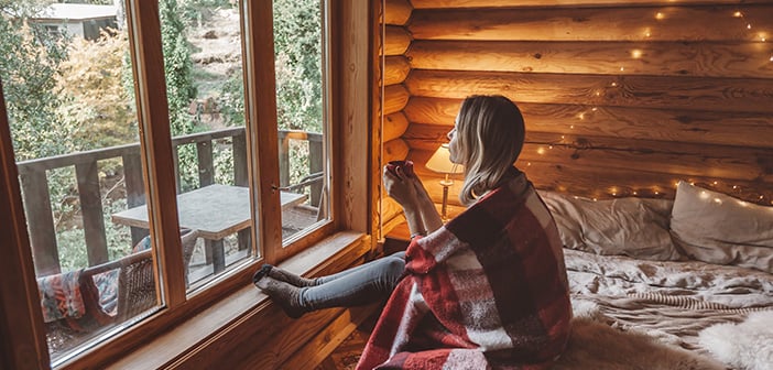 Woman in warm blanket relaxing and drinking morning coffee on cozy bed in log cabin in winter
