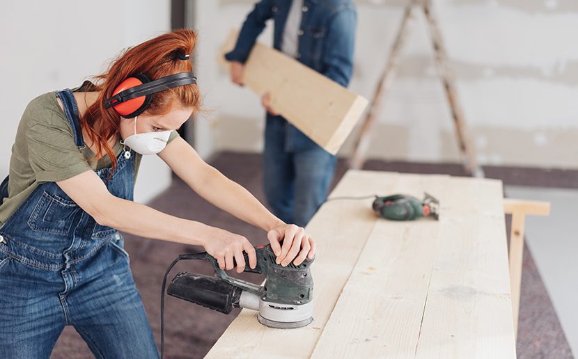 young woman sanding planks of wood with an orbital sander in an unfinished room during DIY renovations
