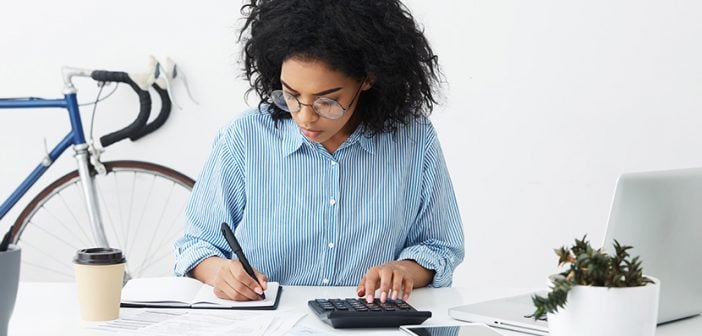 Portrait of attractive hardworking businesswoman with Afro hairstyle busy doing paperwork at office desk, working through finances, using calculator and making notes in her notebook with pen