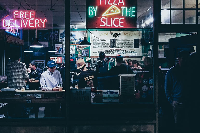 pizza restaurant storefront view through window at night with neon pizza by the slice sign